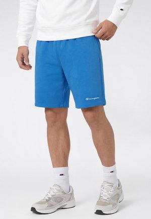 Mężczyzna ubrany w niebieskie szorty Champion, biały sweter, białe skarpetki Champion oraz białe buty sportowe na jednolitym białym tle.