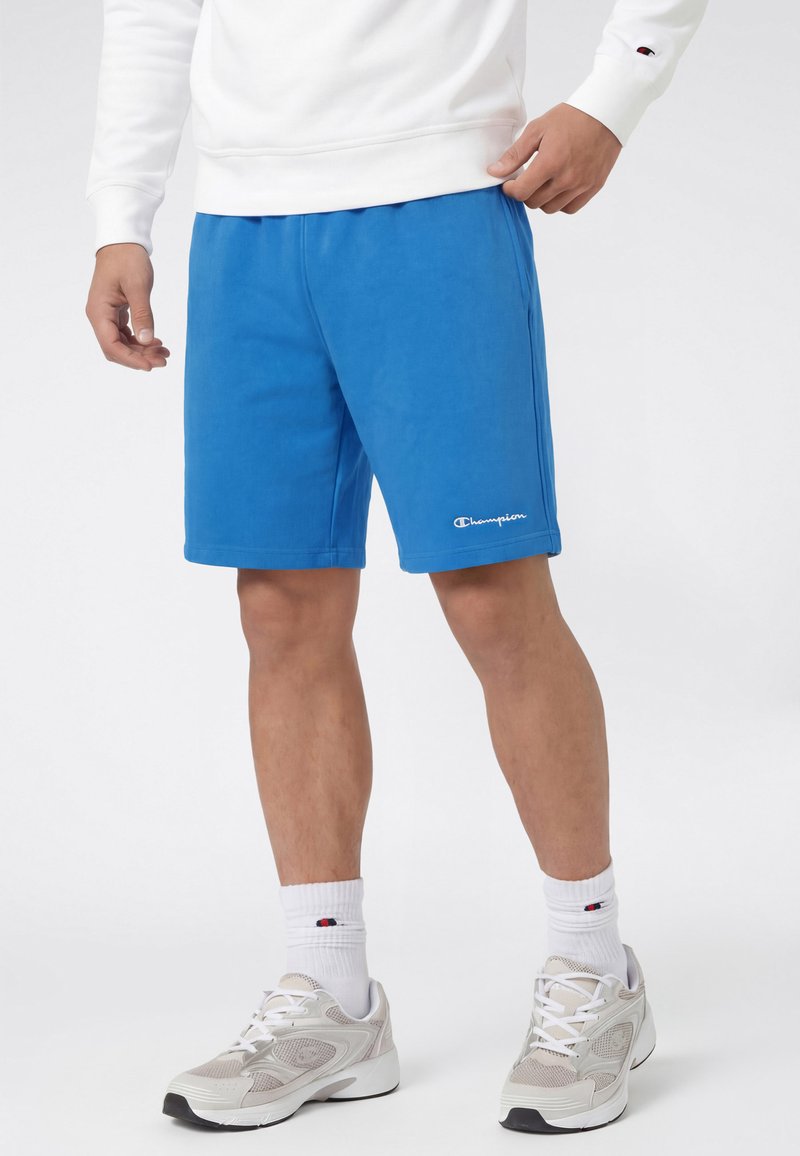 Mężczyzna ubrany w niebieskie szorty Champion, biały sweter, białe skarpetki Champion oraz białe buty sportowe na jednolitym białym tle.