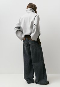 Osoba ubrana w szarą kurtkę z wysokim kołnierzem i szerokie, wyblakłe czarne dżinsy stoi tyłem, trzymając ręce w tylnej kieszeni.