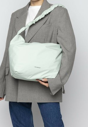 Lichtgroene schoudertas van soepel materiaal, met een geplooide schouderband en subtiele branding. Draagt een grijze oversized blazer.