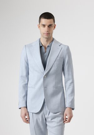 Hombre con cabello corto y oscuro, vestido con un traje azul claro sobre una camisa polo gris, posando frente a un fondo gris liso.