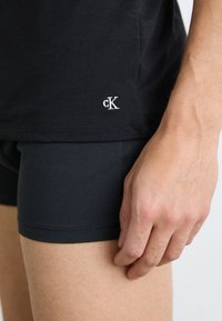 Schwarzes Baumwoll-T-Shirt mit weißem gesticktem Logo am Saum, kombiniert mit grauen dehnbaren Boxershorts, wobei eine Hand auf dem Oberschenkel ruht.
