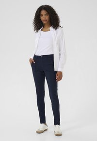 Navy pants in a form-fitting design with front pockets, paired with a white tank top and an open white button-up shirt. Styled with white sneakers.
