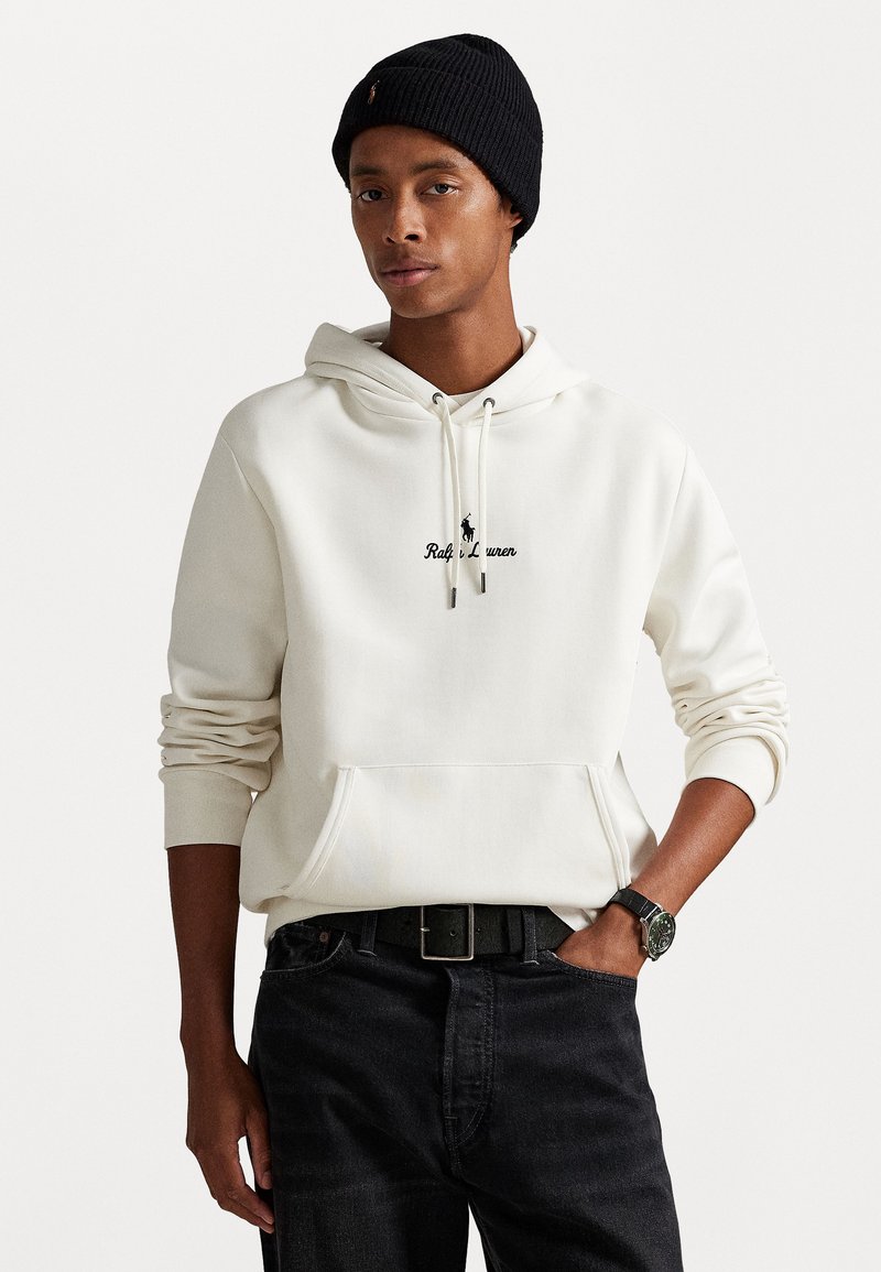 Sweatshirt branca com bolso canguru e logotipo bordado. Combinada com jeans escuros e um gorro preto com nervuras. Relógio complementa o look.