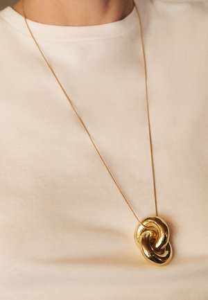 Dunne gouden ketting met een grote, glanzende, abstracte, verweven lus als hanger, gedragen over een beige trui met ronde hals.