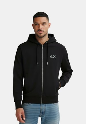 Felpa nera con zip e cappuccio, dotata di un piccolo logo bianco sul petto, texture liscia e polsini a coste. Indossata con jeans blu.