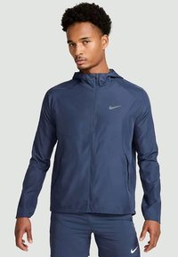 Bărbat îmbrăcat într-o jachetă cu fermoar Nike bleu marin și pantaloni scurți asortați, stând pe un fundal gri deschis uniform.