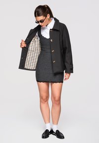 Femme portant des lunettes de soleil, une chemise blanche, une robe grise à rayures fines et un manteau noir dévoilant une doublure à carreaux, des mocassins noirs et des chaussettes blanches.