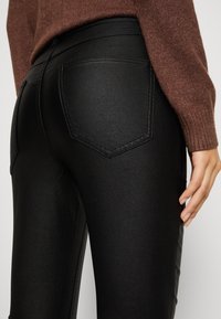 Jean noir taille haute en tissu brillant, avec deux poches arrière aux bords cousus et un panneau latéral texturé.