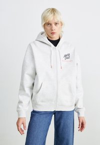 Sweat à capuche gris zippé, arborant un logo en noir et rose sur le devant. Il possède deux poches latérales et des poignets et un ourlet côtelés.