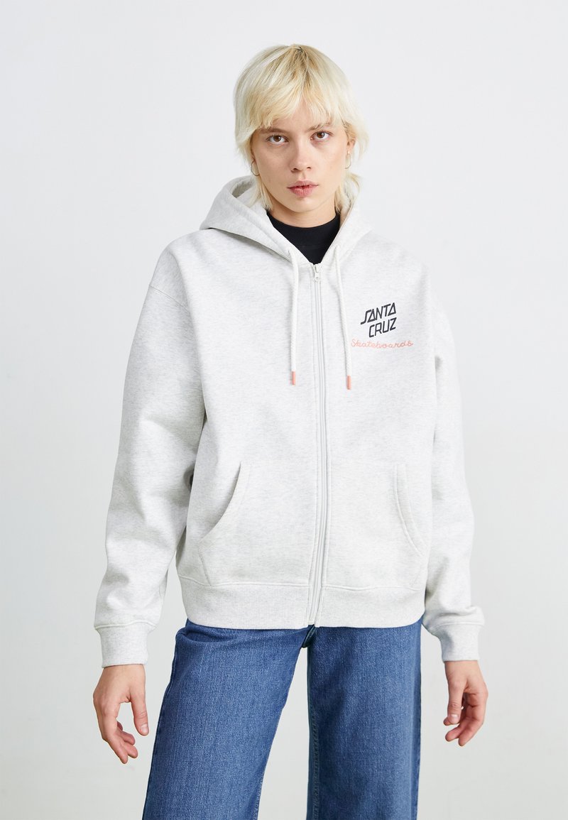 Sweat à capuche gris zippé, arborant un logo en noir et rose sur le devant. Il possède deux poches latérales et des poignets et un ourlet côtelés.