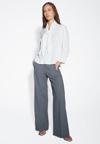 Blusa bianca con scollo annodato, maniche lunghe e vestibilità ampia. Pantaloni grigi a gamba larga con pieghe e tasche. Tessuto semplice e liscio.
