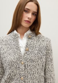 Cárdigan de punto en gris y blanco con cuello de canalé, que presenta tres botones en tono dorado y un patrón texturizado. Llevado sobre una camisa blanca.