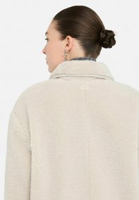 Donna con i capelli raccolti in un chignon intrecciato, indossa orecchini a cerchio in argento e un cappotto beige peloso, vista di spalle su uno sfondo bianco.