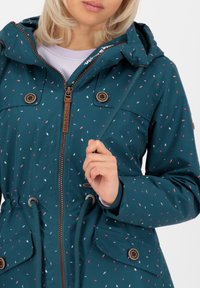 Giacca impermeabile teal con cappuccio, dotata di zip, vita regolabile con cordino e motivo a macchie multicolori. Include tasche anteriori e dettagli decorativi.
