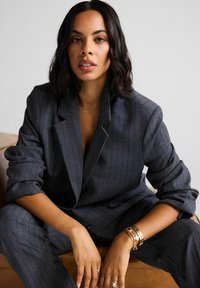 Traje gris a rayas con un blazer de solapa ancha y pantalones ajustados. Las manos del modelo muestran múltiples accesorios dorados y rojos; pose suave y sentado.