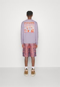 Persona con capelli intrecciati che indossa un felpa grafica viola, pantaloni corti rossi a motivi, calze bianche e stivali beige, in piedi contro un muro bianco.