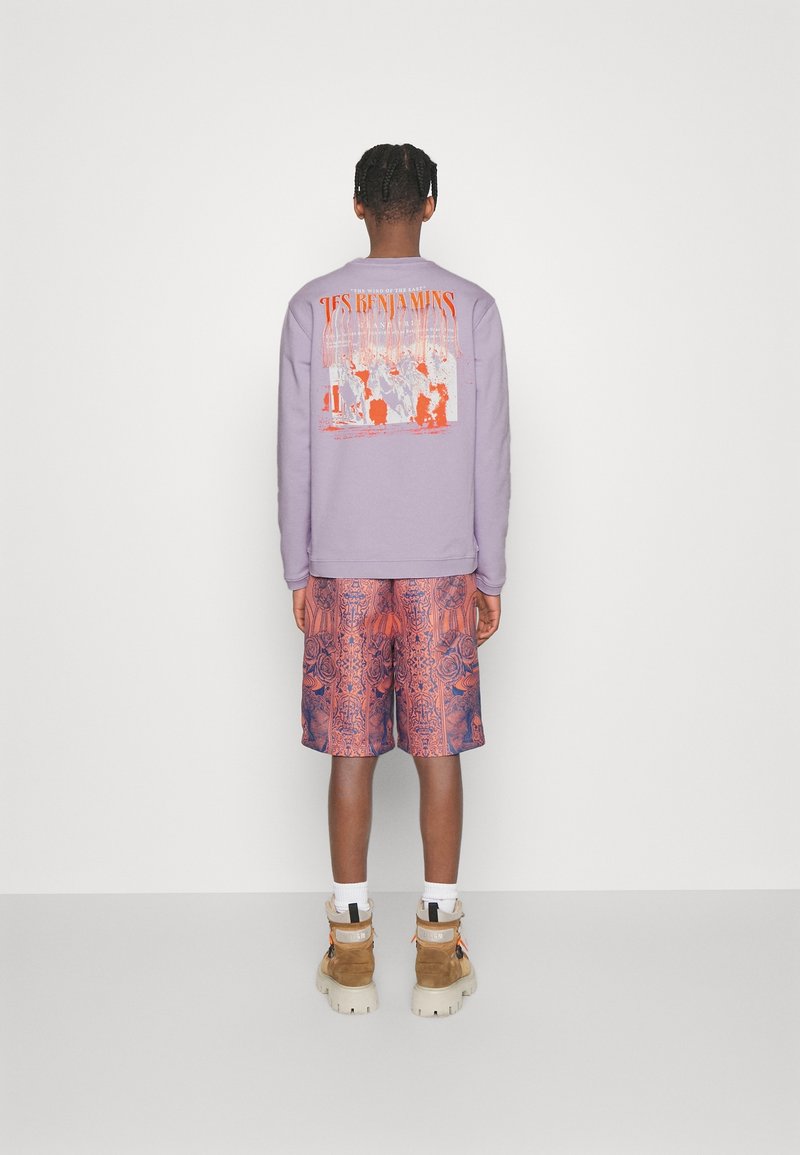 Persona con capelli intrecciati che indossa un felpa grafica viola, pantaloni corti rossi a motivi, calze bianche e stivali beige, in piedi contro un muro bianco.