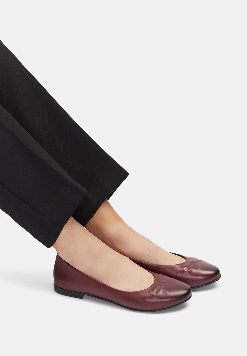 Stopy w ciemnoczerwonych skórzanych baletkach, zestawione z czarnymi spodniami, na białym tle.