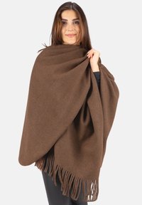 Frau, die einen braunen Fransenschal aus Wolle über schwarzer Kleidung trägt, hält den Schal nahe ihrer Schulter, neutraler Hintergrund.
