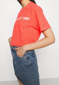 Korálově oranžové bavlněné tričko s krátkými rukávy, s nápisem "TOMMY" v bílé barvě. V kombinaci s džínovou sukní, která ukazuje ležérní střih.