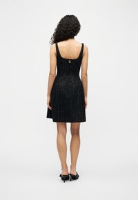 Γυναίκα μ ε κατσαρά μαλλιά που φοράει μαύρο αμάνικο φόρεμα με διακριτικό χρυσό πλέγμα και μαύρα ψηλοτάκουνα παπούτσια, όρθια και γυρισμένη προς τα πίσω.