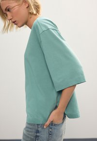 Tricou verde mentă cu mâneci scurte, croială lejeră, textură moale și crăpături laterale, asortat cu blugi de culoare albastru deschis.