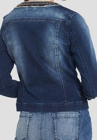 Vista posteriore di una persona che indossa una giacca di jeans blu scuro sbiadita su jeans di denim azzurro contro uno sfondo semplice.