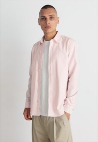 Lys rosa, vertikalt stripete langermet skjorte med knapper, lagdelt over en hvit T-skjorte, kombinert med lyse beige bukser.