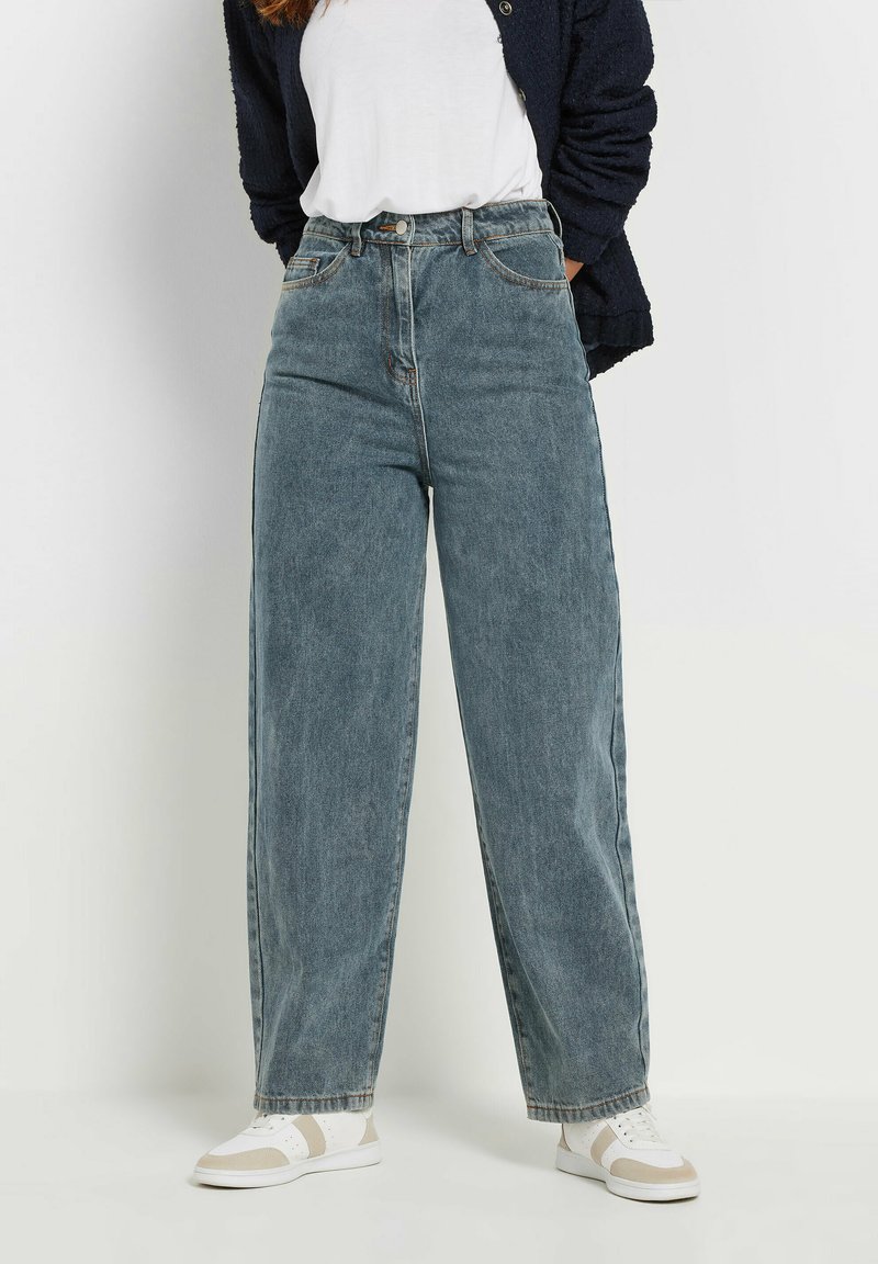 Hoge taille wijde spijkerbroek in vervaagd blauw, met een knoopsluiting, vijf zakken en een onafgewerkte zoom, gecombineerd met witte sneakers.