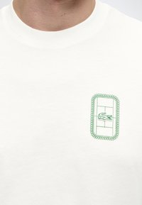 Λευκό T-shirt με μικρή πράσινη κεντημένη εικόνα γηπέδου τένις και λογότυπο κροκόδειλου στην αριστερή πλευρά του στήθους.