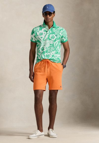 Άνδρας που φοράει πράσινο πουκάμισο πόλο με λουλουδάτο σχέδιο, πορτοκαλί σορτς, λευκά αθλητικά παπούτσια, μπλε καπέλο και γυαλιά ηλίου, στέκεται με το ένα χέρι στην τσέπη μπροστά σε ουδέτερο φόντο.