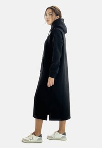 Vestido sudadera negro de gran tamaño con mangas largas, aberturas laterales y un bolsillo tipo canguro. Lleva zapatillas blancas con detalles en gris.