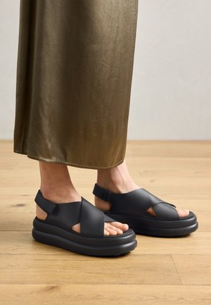 Zwarte platform sandalen met kruisbanden en open hielontwerp, gemaakt van soepel materiaal, gedragen met een vloeiende olijfgroene rok op een houten vloer.