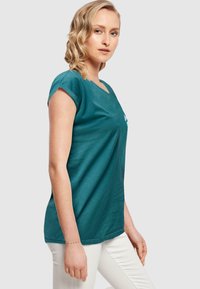Türkisfarbenes V-Ausschnitt-T-Shirt mit kurzen umgeschlagenen Ärmeln, aus weichem Stoff gefertigt, mit einem dezenten Frontdesign, kombiniert mit weißen Hosen.
