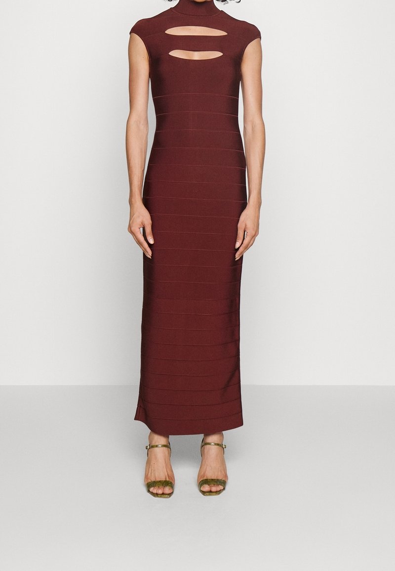 Robe ajustée sans manches bordeaux avec un col haut et deux découpes horizontales sur la poitrine, portée avec des sandales vertes à bride à la cheville.