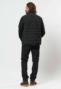 Homme aux cheveux bouclés portant une veste noire matelassée, un jean noir et des chaussures marron, debout de dos sur un fond clair uni.