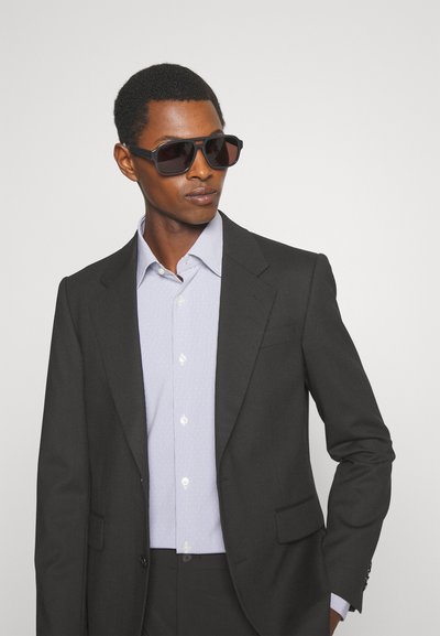 Férfi fekete öltönyben, világos csíkozott ingben és nagy sötét napszemüvegben, egy sima világos háttér előtt áll.