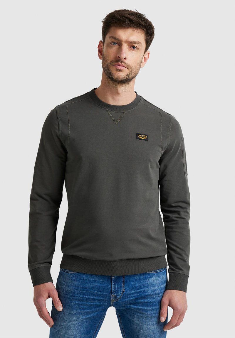 Uomo che indossa una felpa grigio scuro a maniche lunghe con collo rotondo e una piccola toppa sul petto e jeans blu, in piedi davanti a uno sfondo semplice.