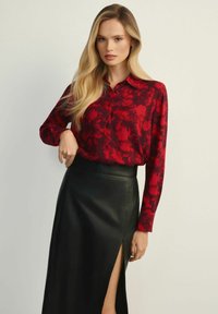 Kobieta z długimi blond włosami, ubrana w czerwoną bluzkę w wzory, włożoną w czarną skórzaną spódnicę z wysokim rozcięciem, stojąca na jednolitym tle.