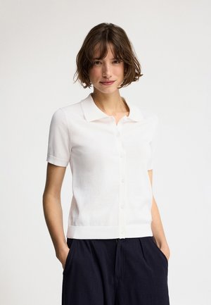 Femme aux cheveux bruns courts portant une chemise blanche à manches courtes boutonnée et un pantalon sombre, les mains dans les poches, fond neutre.