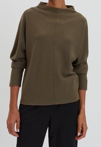 Haut texturé vert olive avec un col montant, des manches trois-quarts et une coupe décontractée, associé à un pantalon noir.