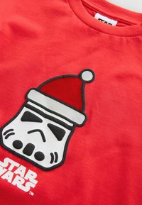 Červené bavlněné tričko s potiskem helmy stormtroopera nosícího vánoční čepici, s černým obrysem a bílými akcenty. Logo umístěné níže.