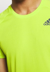 Ljusgul sporttröja gjord av andningsbar, strukturerad tyg. Har en rund halsringning och en mörk logotyp på övre vänstra sidan.