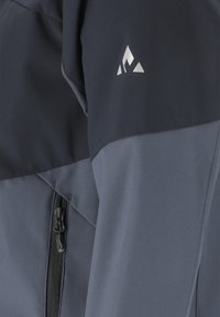 Lähikuva harmaan ja mustan kaksivärisen takin hihasta, jossa on vetoketjullinen tasku ja pieni valkoinen geometrinen logo kyynärpään yläpuolella.