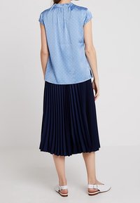 Woman wearing a light blue short-sleeve polka dot blouse, navy pleated midi skirt, and white low-heeled strap shoes, standing facing away.