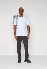 Camiseta blanca con logotipo azul, pantalones oscuros con bolsillos, zapatillas negras y una mochila de un azul claro. Complementado con una gorra beige.