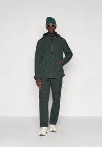 Donkergroene waterdichte jas met een rits aan de voorkant, capuchon en zakken, gecombineerd met bijpassende cargobroek, witte sneakers en een groene beanie.
