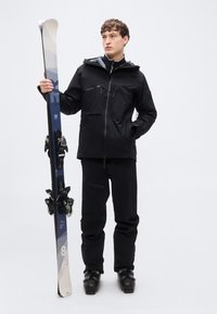 Μαύρο αδιάβροχο σακάκι σκι με τσέπες με φερμουάρ, σε συνδυασμό με μαύρο παντελόνι σκι. Το μοντέλο κρατά μπλε και λευκά σκι με δεσίματα. Μαύρα μπουτάκια σκι.