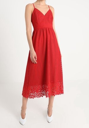 Sleeveless red midi dress with spaghetti straps, V-neck, perforated fabric, and floral cut-out hem, worn with white pointed shoes.